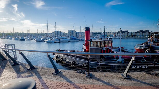 A marina with boats docked, a red boat named 'BARKING' in the foreground, and a cityscape in the background.