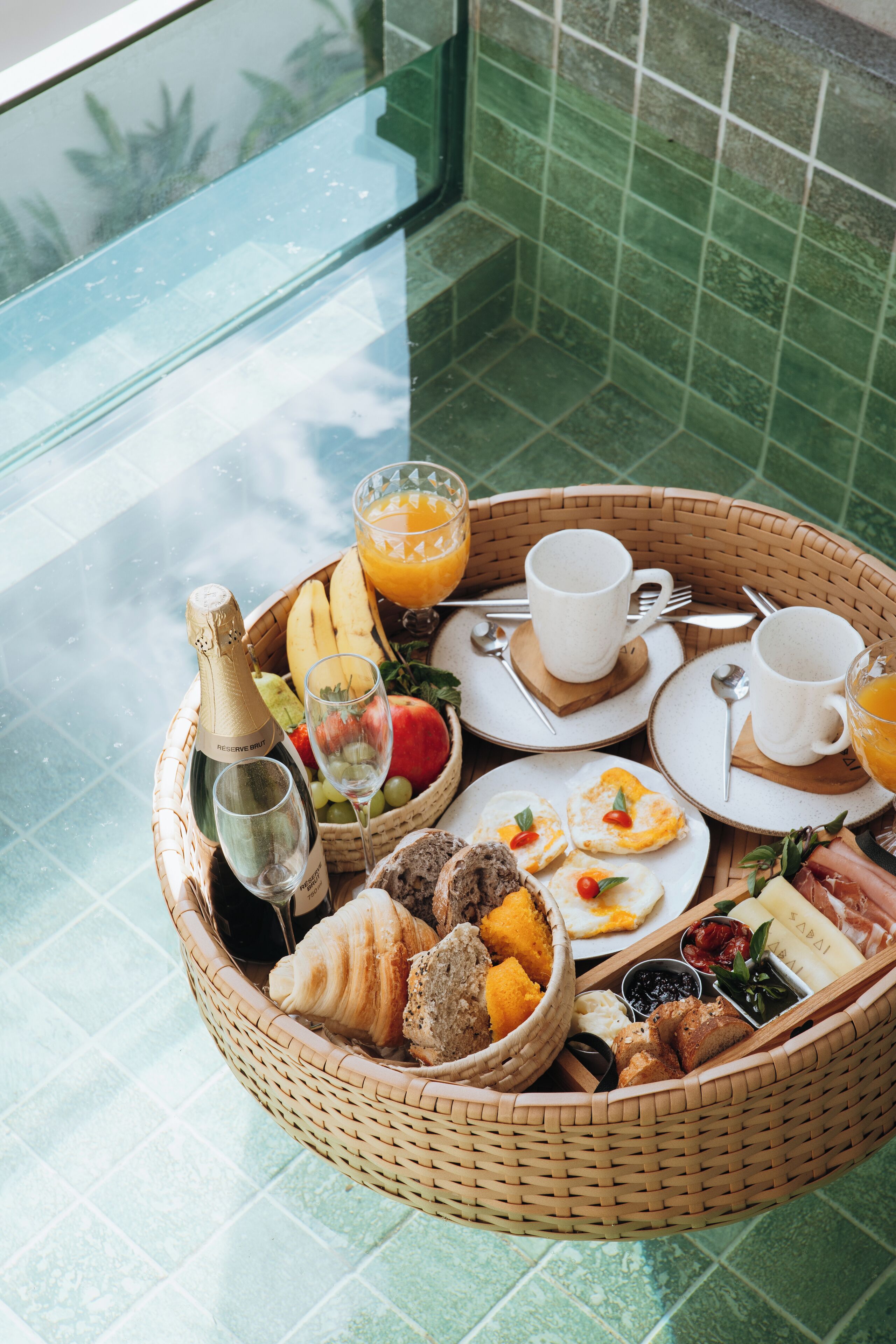 A breakfast tray with a bottle, glasses, fruits, pastries, and a bowl of soup on a glass table.