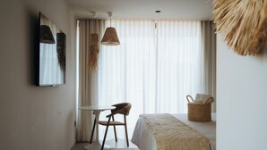 Habitación de hotel con cama, una mesita, una silla, una cesta tejida y un ventanal con cortinas traslúcidas.