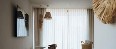 A hotel room with a bed, a small table, a chair, a woven basket, and a large window with sheer curtains.