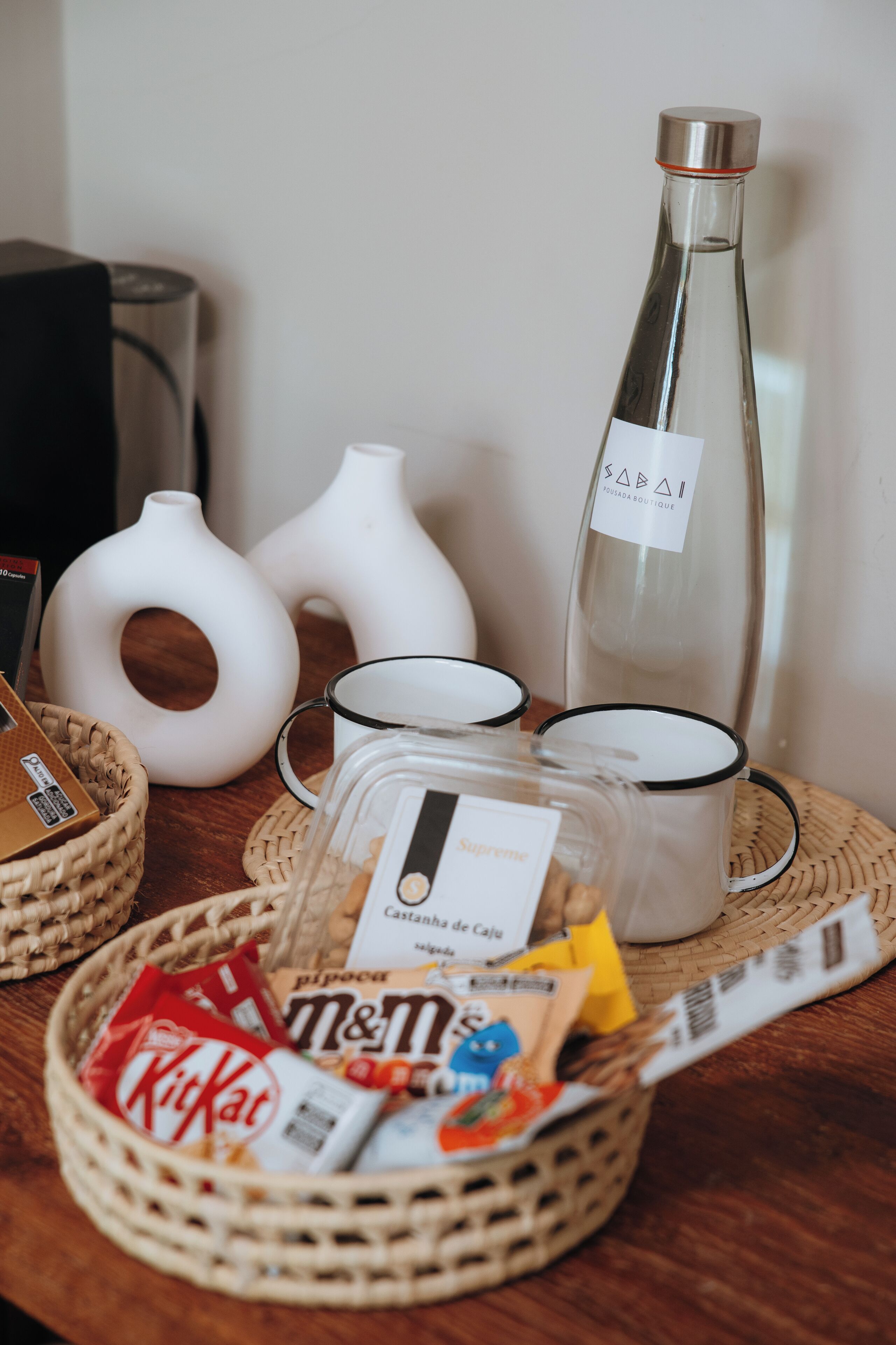 Una mesa con una botella, dos jarrones blancos, una cesta de aperitivos y una bandeja con tazas y una caja etiquetada "Supreme".