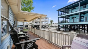 A covered outdoor deck with wooden furniture, a white railing, and a view of a multi-story building with balconies. - Ocean Lakes Z49 | Lakeview 3BR Ground-Level Retreat Comp Golf Cart (Myrtle Beach)
