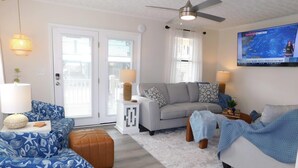 A living room with a sofa, ottoman, and a television displaying a weather forecast. - Ocean Lakes Z49 | Lakeview 3BR Ground-Level Retreat Comp Golf Cart (Myrtle Beach)
