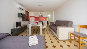 Quarto de hotel pequeno com cama, sofá, mesa de jantar com cadeiras, televisão e cozinha compacta.