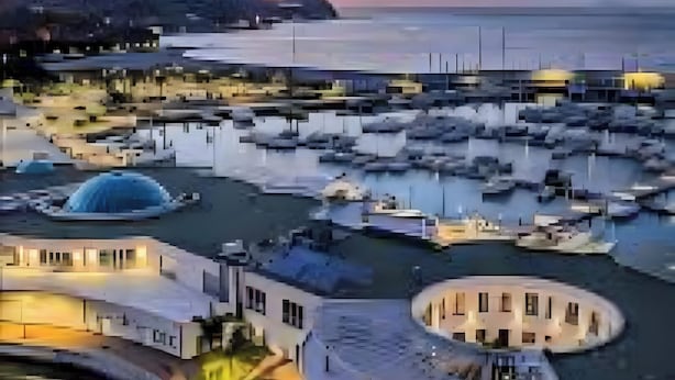 Uma marina costeira com barcos, um edifício em formato de cúpula e um calçadão à beira-mar.