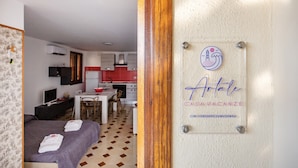 Quarto de hotel com cama, escrivaninha, cadeira, televisão e área de cozinha ao fundo.