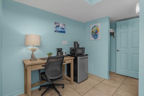 A room with a desk, chair, lamp, coffee maker, microwave, and artwork on the walls.