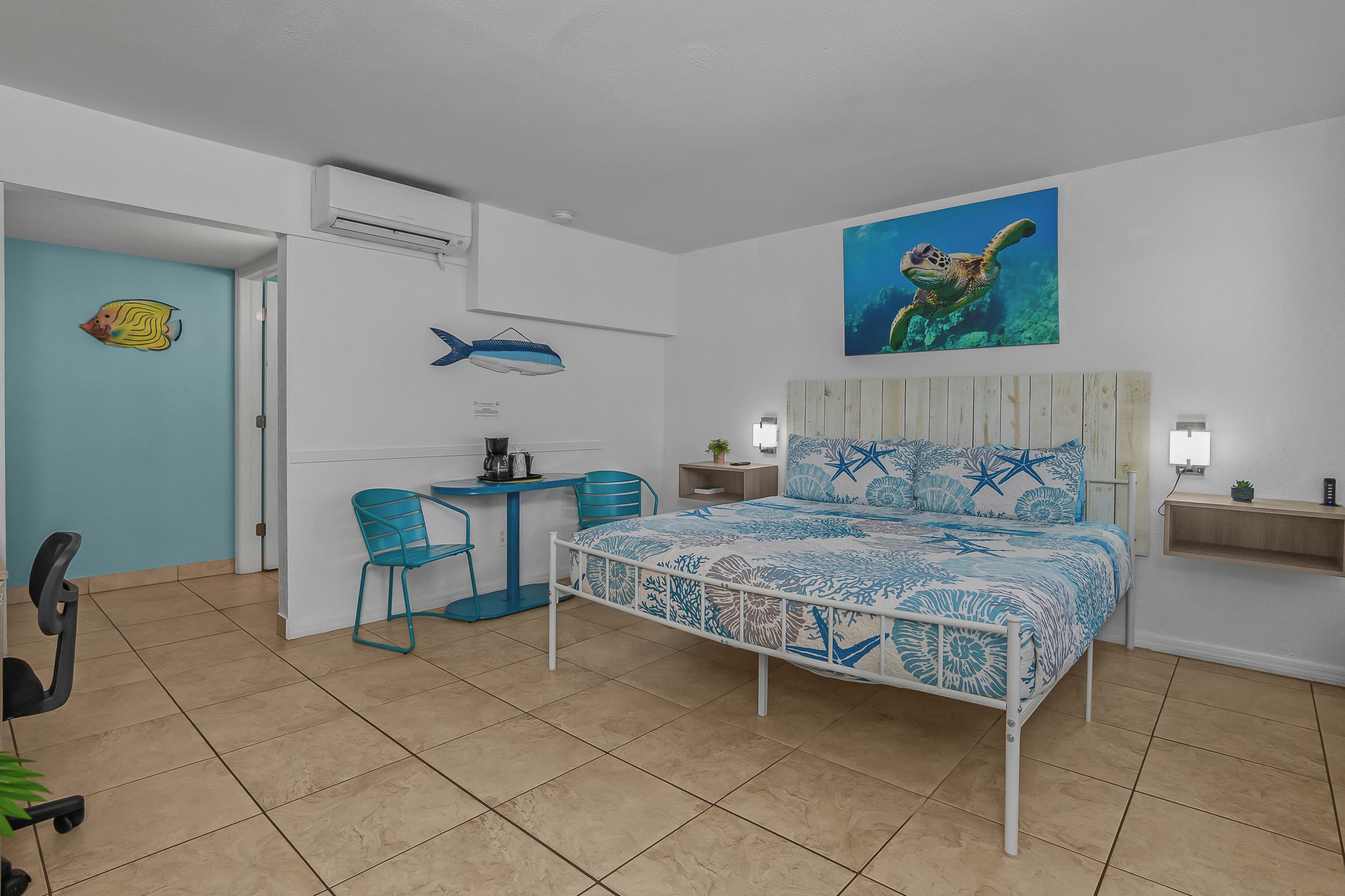 A bedroom with a bed, a desk, chairs, and a wall with a fish and a turtle painting.