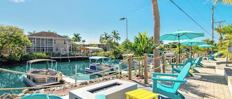 Una zona frente al agua con barcos amarrados, mesas al aire libre y un edificio al fondo.