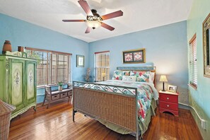 Ein Schlafzimmer mit einem Bett, einem grünen Schrank, einem Korbstuhl, einem Deckenventilator und einem Fenster mit Jalousien.