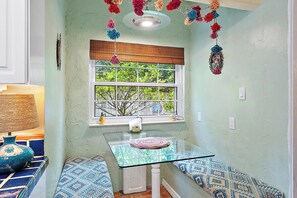 Une petite pièce avec une table en verre, des coussins à motifs, une fenêtre donnant sur un paysage verdoyant et des pompons décoratifs.