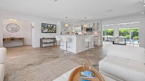 A modern kitchen with white cabinetry, a central island with bar stools, and a dining area with a view of greenery. - Bimini Breeze - Updated Island Retreat with Pool & Beach Access (West Palm Beach)