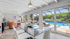 A spacious room with a glass dining table, white chairs, a sofa, and a large window overlooking a swimming pool. - Bimini Breeze - Updated Island Retreat with Pool & Beach Access (West Palm Beach)