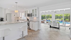 A modern kitchen with white cabinetry, a granite countertop, and a built-in microwave. There is a large window overlooking a pool area with outdoor seating. - Bimini Breeze - Updated Island Retreat with Pool & Beach Access (West Palm Beach)