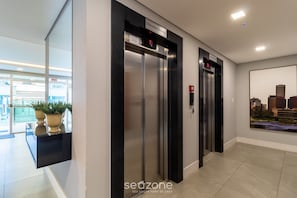 A modern elevator area with two elevators, a digital display, and a cityscape view. - Apt w/ Swimming Pool Well Located Blumenau Neb0103 (Blumenau)