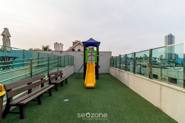 A rooftop playground with a slide, benches, and a glass railing. - Apt w/ Swimming Pool Well Located Blumenau Neb0103 (Blumenau)