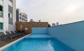 Una piscina con lettini e un muro in legno, circondata da un edificio moderno.