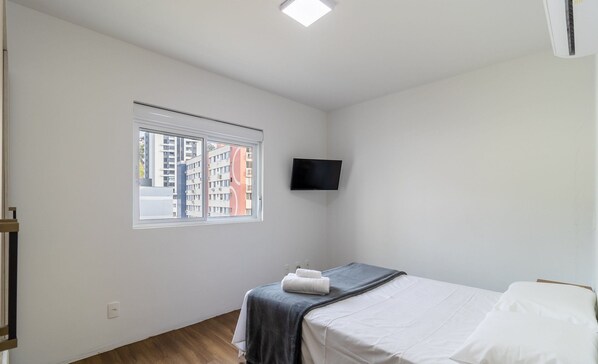 Un dormitorio con cama, ventana, televisor de pantalla plana y suelo de madera.
