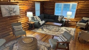 A cozy log cabin interior with a sectional sofa, armchairs, a coffee table, and a large woven rug.