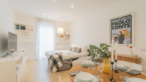 A modern, well-lit room with a bed, a dining table set for two, a TV, and a bookshelf.