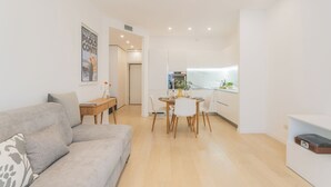 A modern living room with a sofa, a dining table with chairs, and a kitchen area with cabinets and a window.