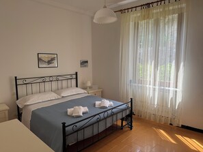 Ein ordentlich bezogenes Bett mit blauer Bettdecke und weißen Kissen, ein Nachttisch mit Lampe und ein Fenster mit transparenten Vorhängen.