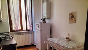 Eine kleine Küche mit einem Gaskochfeld, einem weißen Kühlschrank, einem Tisch mit Blumenmuster und einem Fenster mit Spitzenvorhängen.