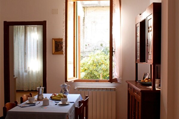 A dining area with a table set for two, a radiator, a window with a view of greenery, and a cabinet with fruit.