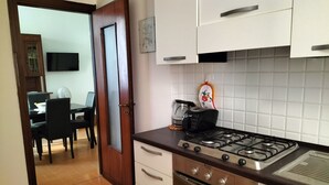 Eine Küche mit Gasherd, Backofen und Kaffeemaschine. Durch eine offene Tür ist ein Essbereich mit Tisch und Stühlen zu sehen.
