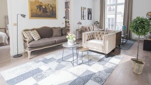 A living room with a beige sofa, a white armchair, a small round table with a vase of flowers, and a large patterned rug.