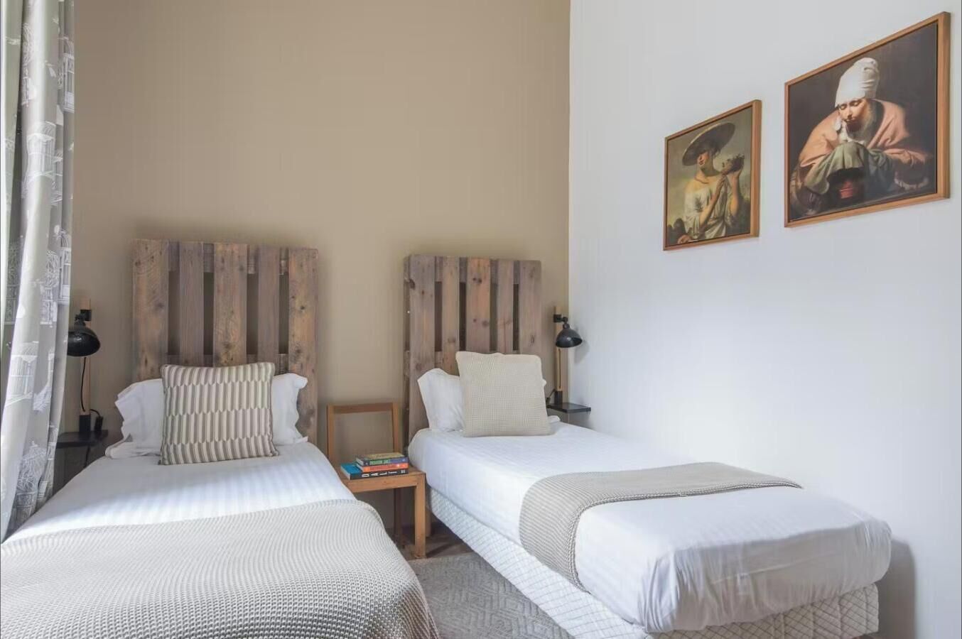 A room with two beds, each with a wooden headboard, white bedding, and a small bedside table with a lamp. There are two framed artworks on the wall.