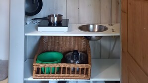A compact kitchen with a sink, a scale, a pan, a whisk, a green bowl, a wicker basket, and a suitcase above.