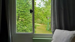 A room with a bed, a window with curtains, and a view of greenery outside.