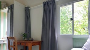A room with a wooden desk, a chair, a window with curtains, and a bed with a green blanket.