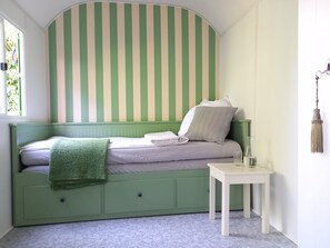 A room with a green-striped headboard, a bed with white and gray bedding, a green blanket, a small white table with a lamp, and a window with greenery outside.