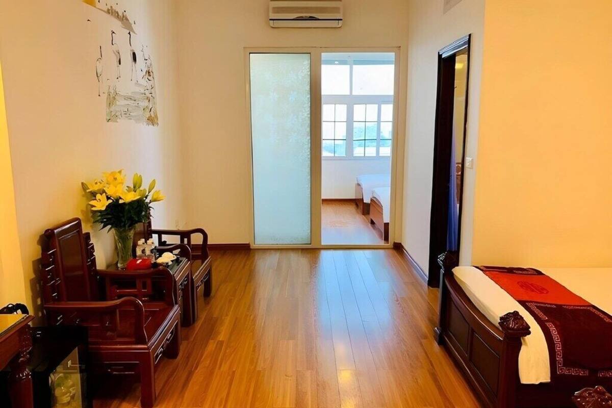 A room with wooden flooring, a bed with a red bedspread, a wooden chair, a small table, and a vase with flowers.