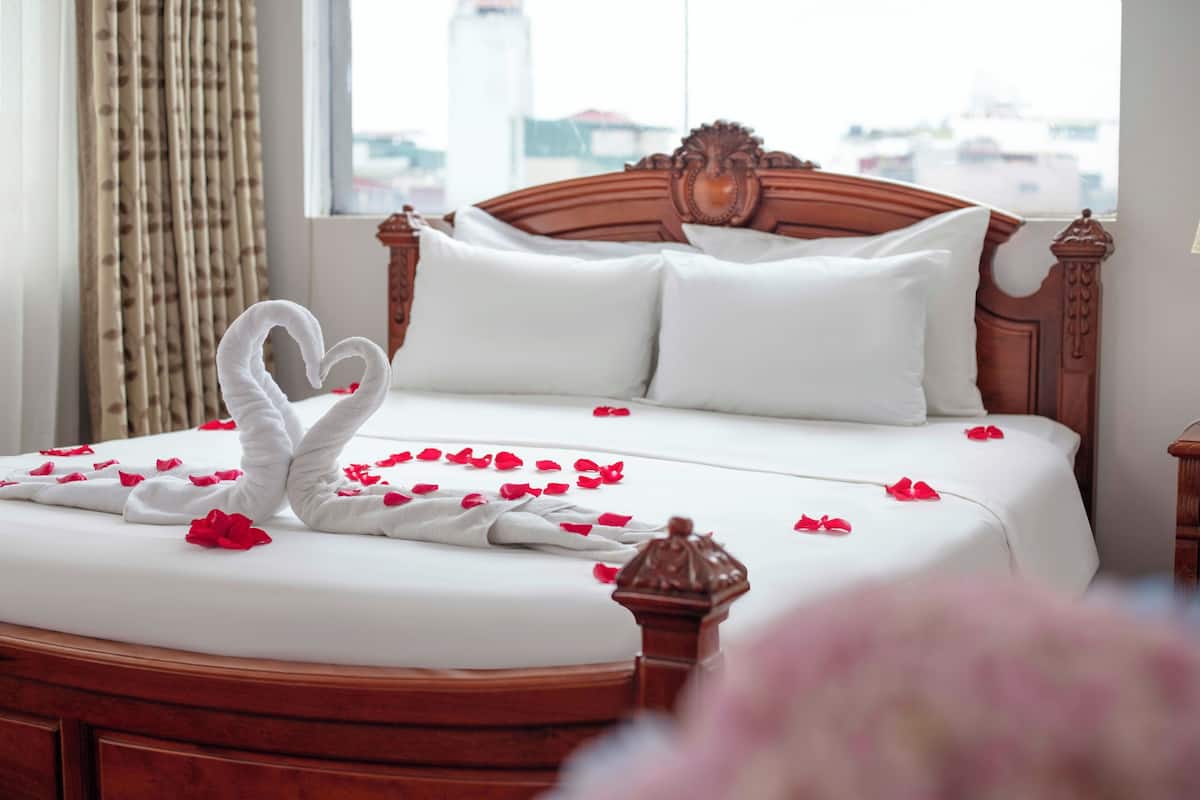 A neatly made bed with a heart-shaped towel and rose petals, a wooden headboard, and a view of buildings through a window.