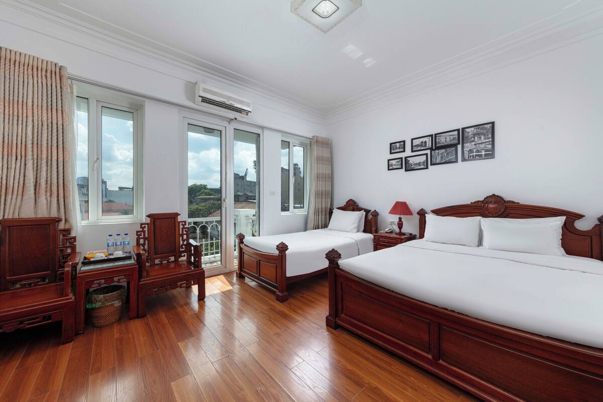 A hotel room with two beds, a wooden desk, a chair, a small table with bottles, and a window with curtains.
