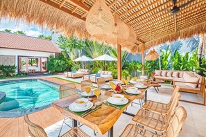 Uma área de refeições à beira da piscina, com uma mesa posta para dois, rodeada por cadeiras de vime, um telhado de palha e vista para a piscina e vegetação.