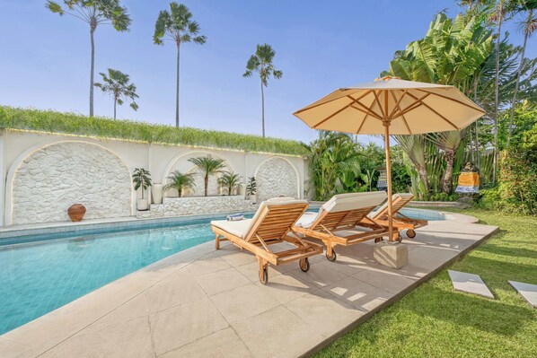 A pool area with two lounge chairs and a large umbrella, surrounded by palm trees and a stone wall. - Villa Manhattan - Luxurious 8-BDR Villa, huge pool (Kecamatan Kuta Utara)