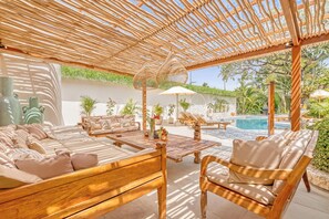 An outdoor lounge area with wooden furniture, a pool, and a pergola providing shade.