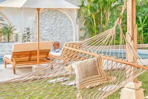 A hammock with a cushion, a wooden lounge chair, and a stone wall in the background. - Villa Manhattan - Luxurious 8-BDR Villa, huge pool (Kecamatan Kuta Utara)