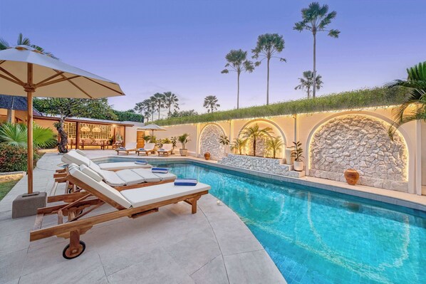 A swimming pool with lounge chairs and umbrellas, surrounded by palm trees and a stone wall.