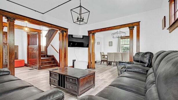 Amplia sala de estar con sofá de cuero negro, piso de madera, una escalera y un comedor con mesa y sillas.