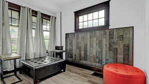 Una habitación con piso de madera, un pouf negro, un pouf rojo, una mesita y una ventana con cortinas.