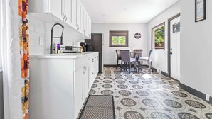 Un couloir étroit, avec un coin cuisine d’un côté, comprenant des meubles blancs, un évier et un réfrigérateur. Au centre du couloir, on trouve une table à manger avec des chaises, et une fenêtre donnant sur un paysage verdoyant.