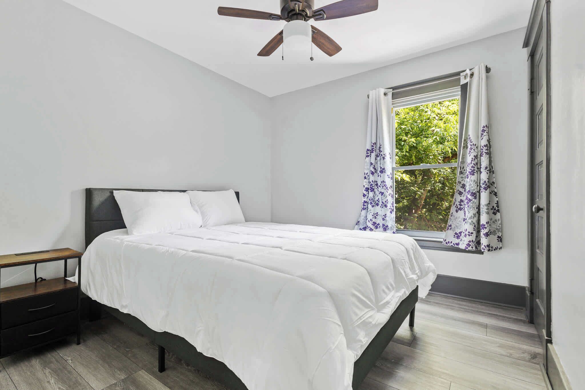 A bedroom with a bed, a nightstand, a ceiling fan, and a window with curtains.
