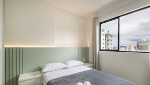 Una habitación de hotel con cama, mesita de noche, ventana con vista a la ciudad y una pared verde.