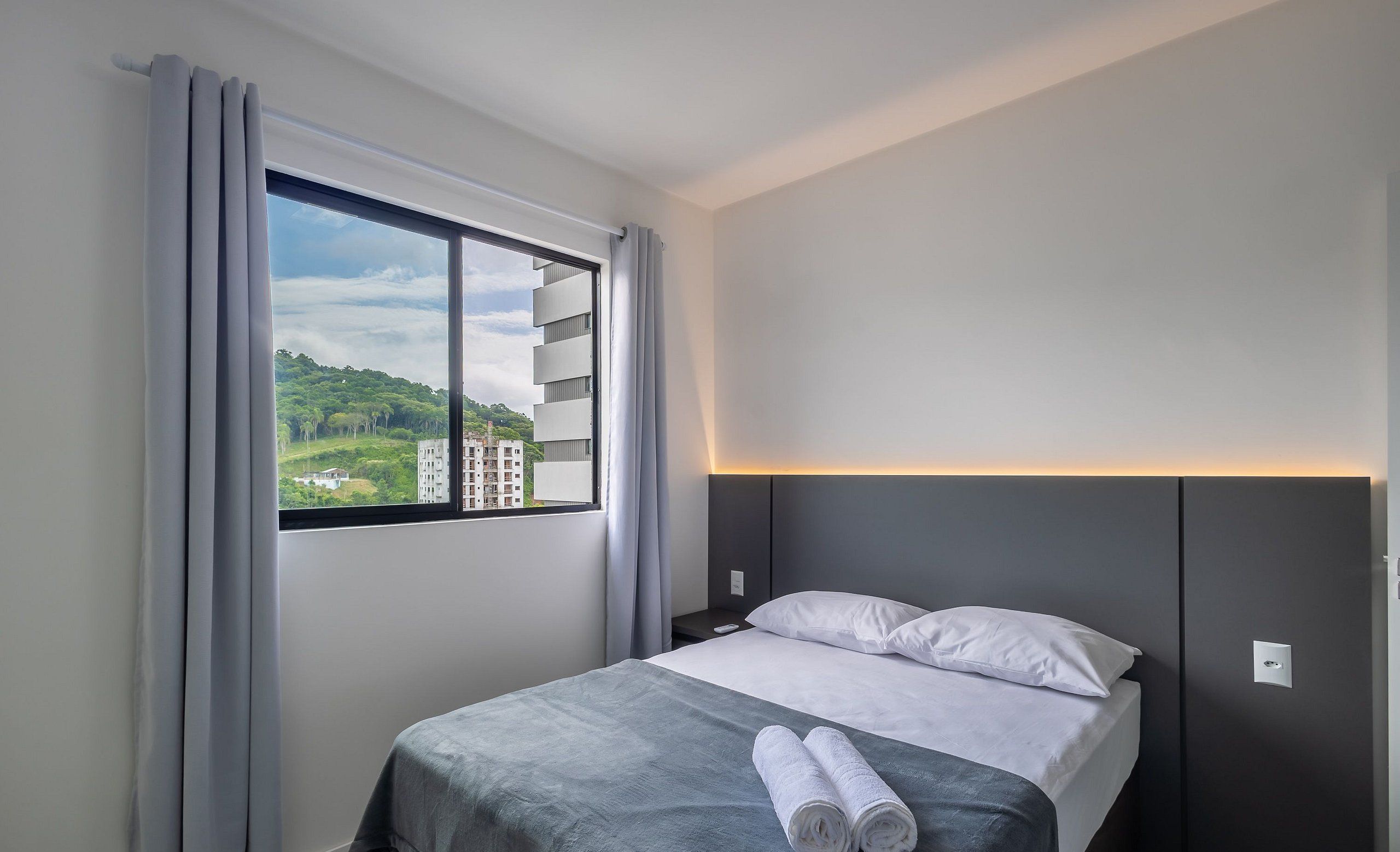 A hotel room with a bed, a nightstand, a window with curtains, and a view of greenery and buildings outside.