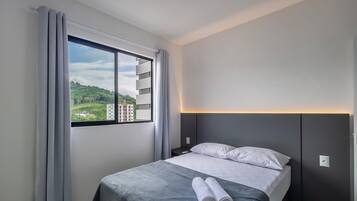 A hotel room with a bed, a nightstand, a window with curtains, and a view of greenery and buildings outside.